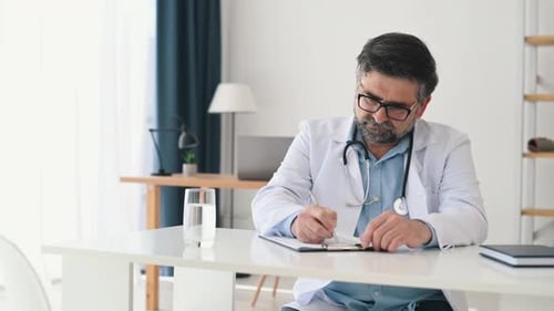 Writing on the document by a pen. Mature male doctor in coat is in the clinic room