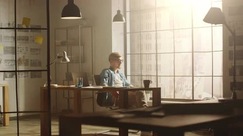 Thoughtful Young Professional Designer Sits at His Desk and Looks onto Big City out of His Windows,