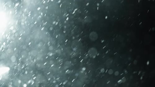 Nahaufnahme wirbelnder Schneeflocken, die nachts von hellem Gegenlicht beleuchtet werden und durch eine Kälte driften