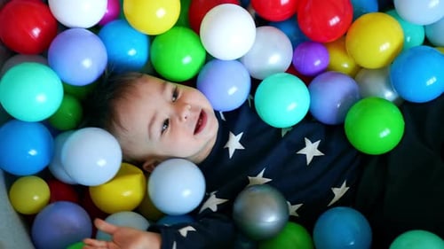 Lovely child lies among the balls inside the dry pool. Happy little kid plays with toys.