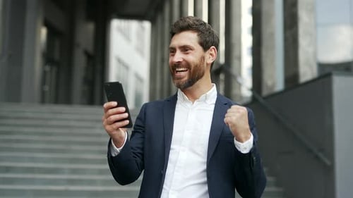 Happy excited businessman in a formal suit received great news on mobile phone standing on street