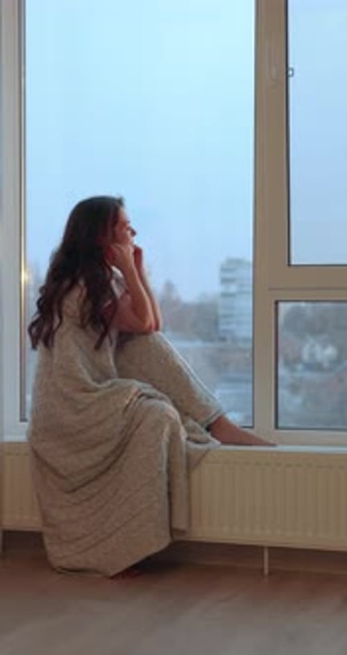 Woman Wrapped in a Blanket Sitting on the Windowsill and Looking Outside