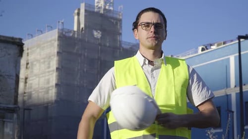 Auf der Baustelle setzt ein junger Bauingenieur mit Brille einen weißen Helm auf und schaut