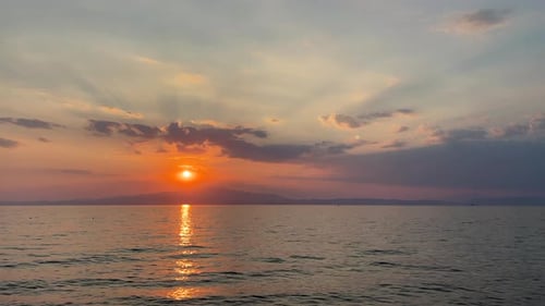 The sun sets on calm waters, casting warm light, with clouds and distant hills under a soft orange s