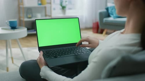 Mujer joven en casa sentada en un sofá trabaja en una computadora portátil con pantalla de maqueta verde. Niña U