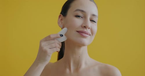 Closeup Portrait of Beautiful Lady Massaging Face with Guasha Scraper Enjoying Skin Care on Yellow