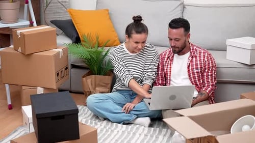 Young Couple Surrounded By Boxes Uses Laptop to Shop for Decor Book Moving Services