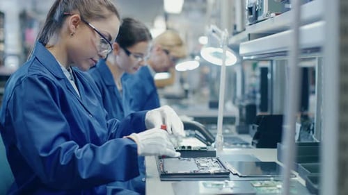 Une travailleuse d'une usine d'électronique en blouse de travail bleue et lunettes de protection assemble un ordinateur portable M