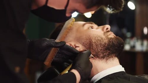 Junger Mann mit stylischer Frisur sitzt und lässt sich im Friseurladen vom Haarschneider den Bart rasieren