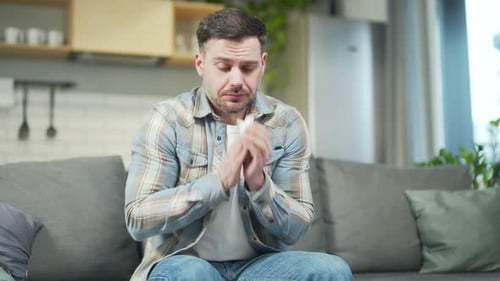 Man Sits on Couch, Sneezes and Rubs Head