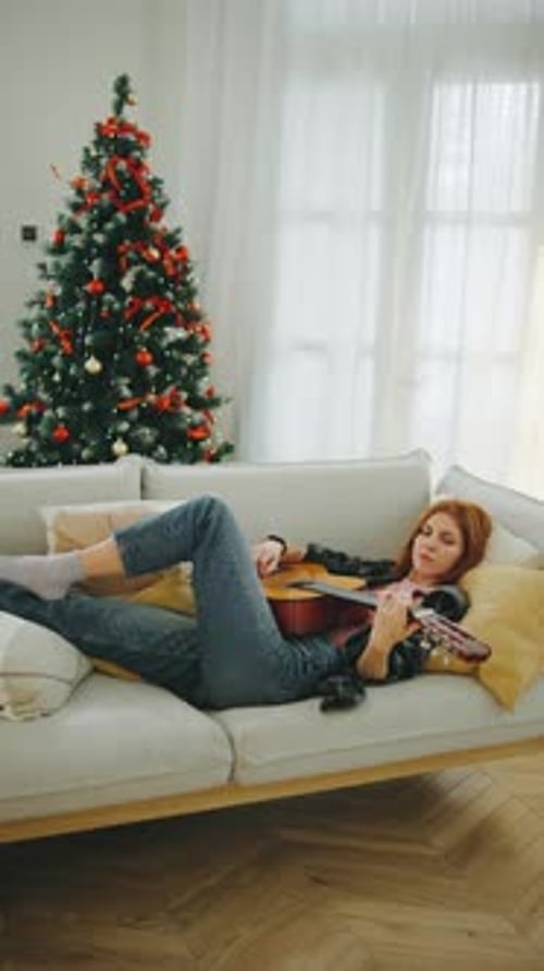 Young Woman Playing Guitar While Relaxing on a Couch Next to a Festive Christmas Tree in a Cozy