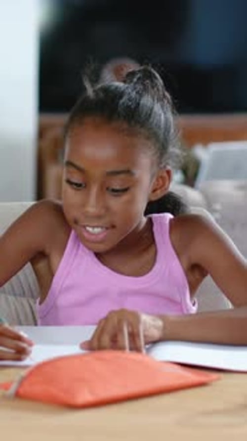 Vertical video: Leaning girl in pink top writing for homework at home, orange pencil case visible