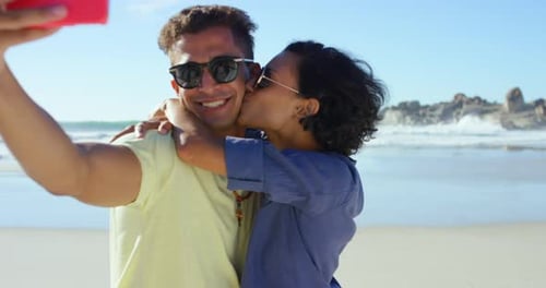 4k video footage of a young couple standing together on the beach and taking selfies on a cellphone
