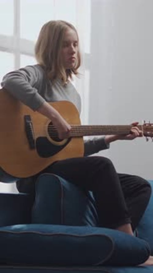 Teenage Girl Practicing Acoustic Guitar on Blue Sofa in Bright Cozy Living Room Focused on Learning