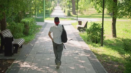 Young Man Walking Sunlit Park Path Student Returning From Class Messenger Pausing By Bench Commuter