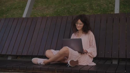 Handheld Close Up of a Curly Caucasian Woman Working on Her Laptop Computer on a Park Bench