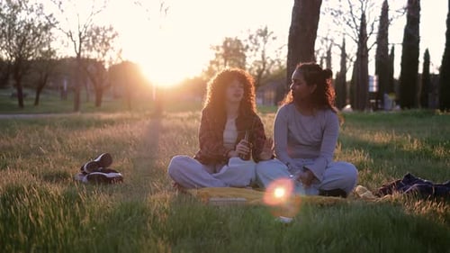 Two young women, likely students, are sitting on a blanket in a park, enjoying a picnic and conversa