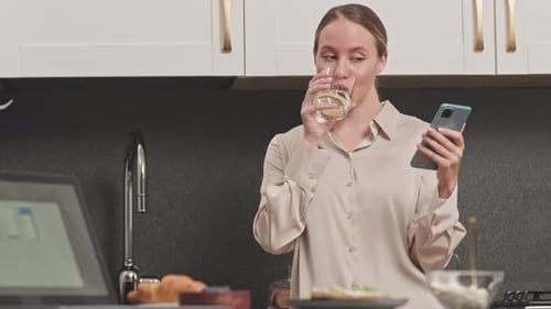 Woman Using Phone While Drinking Water in Kitchen