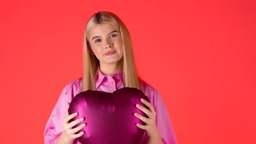 Pretty Blonde Woman Holding Purple Heart Shaped Balloon Against Red Background, Studio Shot