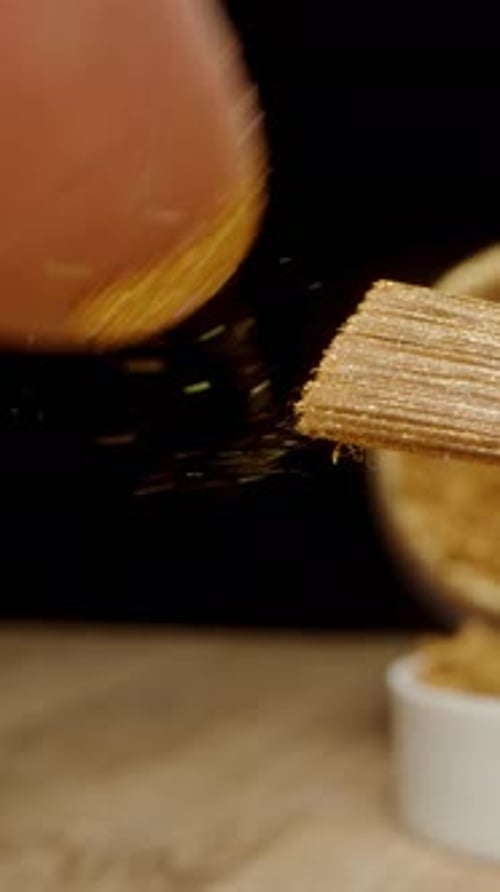 Vertical. a brush with golden particles of powder or dust on it, being flicked by a finger.