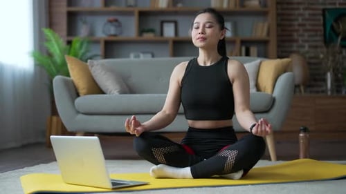 Young Asian woman meditates during an online course on laptop sitting on mat on floor in living room