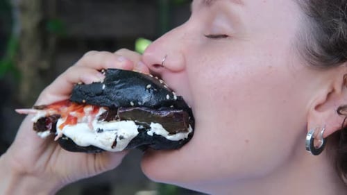Closeup of a Woman Takes a Big Bite From an Appetizing Black Burger with Sauce and Chews Delicious