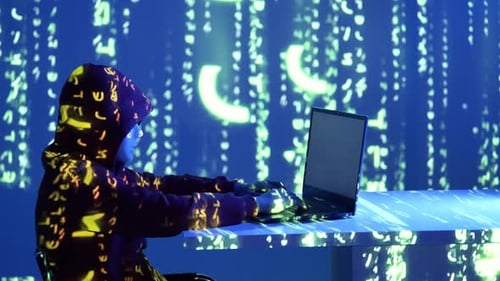 Cyberpunk hacker in hooded sweatshirt coding on laptop with green matrix code on blue digital wall