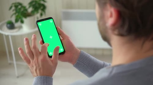 Young Man Sitting at Home Holding Smartphone Green Mockup Screen in Hand Male Person Using Chromakey