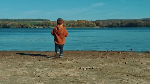 Energetic baby boy picking stones on the river bank. Kid in warm jacket throws stones into water.