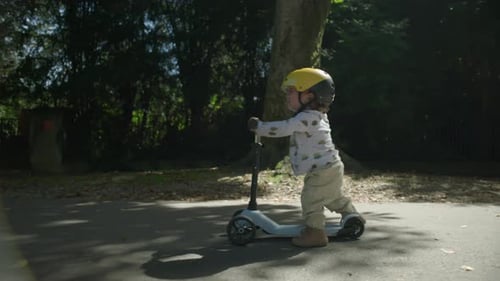 Toddler with yellow helmet walking beside scooter in park, starting to ride with tearful expression