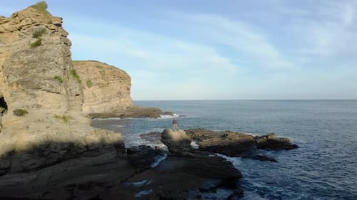 Aerial shot flying over a man standing on top of a rocky outcrop on the edge of the ocean at the bot