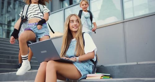 Young beautiful girl uses laptop meets and greets school friends while sitting on stairs near school
