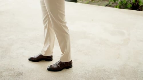 Close-up of a groom in a white suit wearing dark brown leather shoes, walking on a light pavement. Close-up of a groom in a white suit wearing dark brown leather shoes, walking on a light pavement.