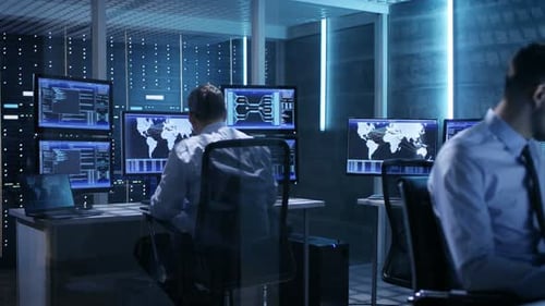 Dos profesionales de TI que trabajan en una sala de monitoreo rodeada de varias pantallas.System Co