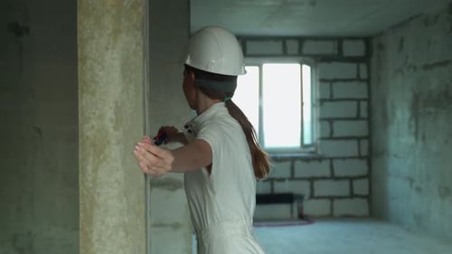 A Focused Handyman in a White Helmet and with a Tape Measure Measures the Wall on the Construction