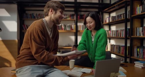 Guy and Girl Students Communicate and Discuss Ideas While Sitting on a Table in the Library
