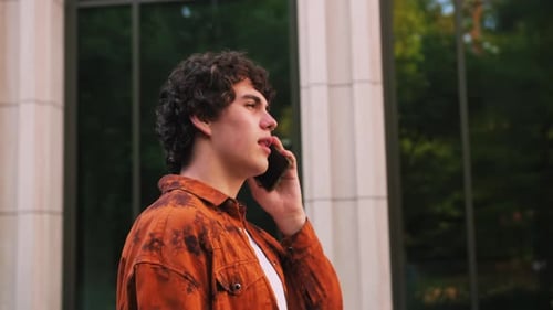 Closeup of Young Man in Orange Shirt Talking on Mobile Phone and Walking Down the Street Modern