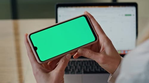 Woman Holds Smartphone with Green Screen by Laptop