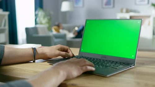 Over the Shoulder Shot of Mans Hands Typing on a Laptop with Green Mock-up Screen. In the Backgroun
