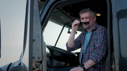 Close Up Portrait of Professional Truck Driver Sits in His Truck and Behind Him Parked Long Haul