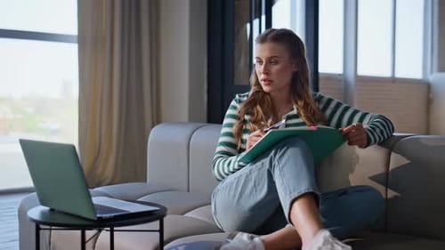 Woman Writes in Notebook on Couch with Laptop