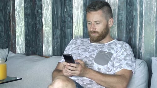 Young Man Using Smartphone and Relaxing on Sofa in Cafe 30s