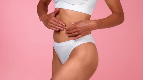 Frau mit mittlerem Hautton hält ihren Bauch fest, während sie weiße Kleidung vor rosafarbenem Hintergrund trägt