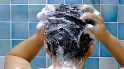 Male hands wash their hair with shampoo and foam against background of blue tiles, rear view.