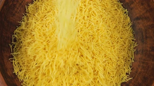 Thin small pasta is poured into a brown ceramic bowl. Top view. Slow motion.