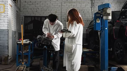 Student Measuring Engine Component While Colleague Records Data in Automotive Workshop