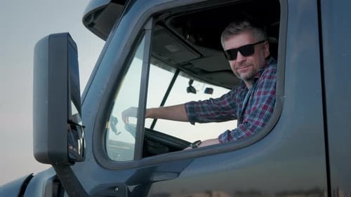 Professional Truck Driver Sits in His Truck and Behind Him Parked Long Haul SemiTruck