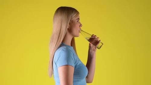 Side View Of Blonde Woman Drinking Water From Glass Bottle, Studio Portrait