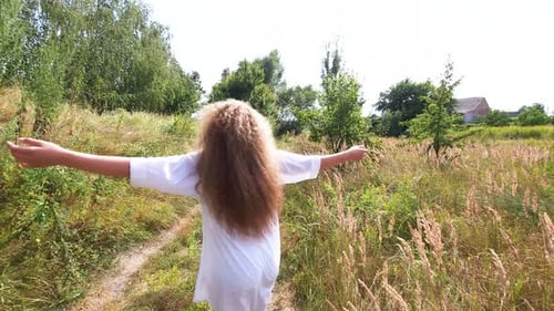 A blond girl with long hair in a white shirt is spinning in the middle of a meadow with field herbs
