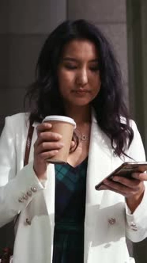 Vertical video: Sipping woman in blazer drinking from paper cup, scrolling phone outside stone door